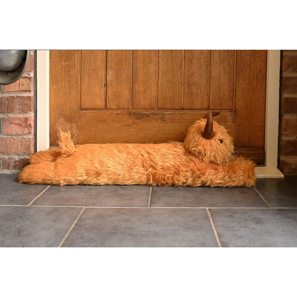 Orange plush Highland cow draught excluder under door opening, long body and horns shown.
