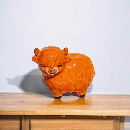 Glossy orange Highland cow figurine on a wooden shelf, full body profile