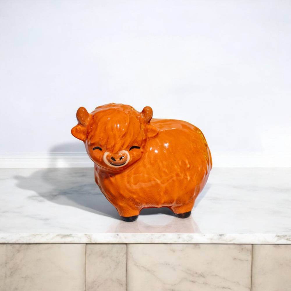 Glossy orange Highland cow figurine standing on a light stone surface, side view