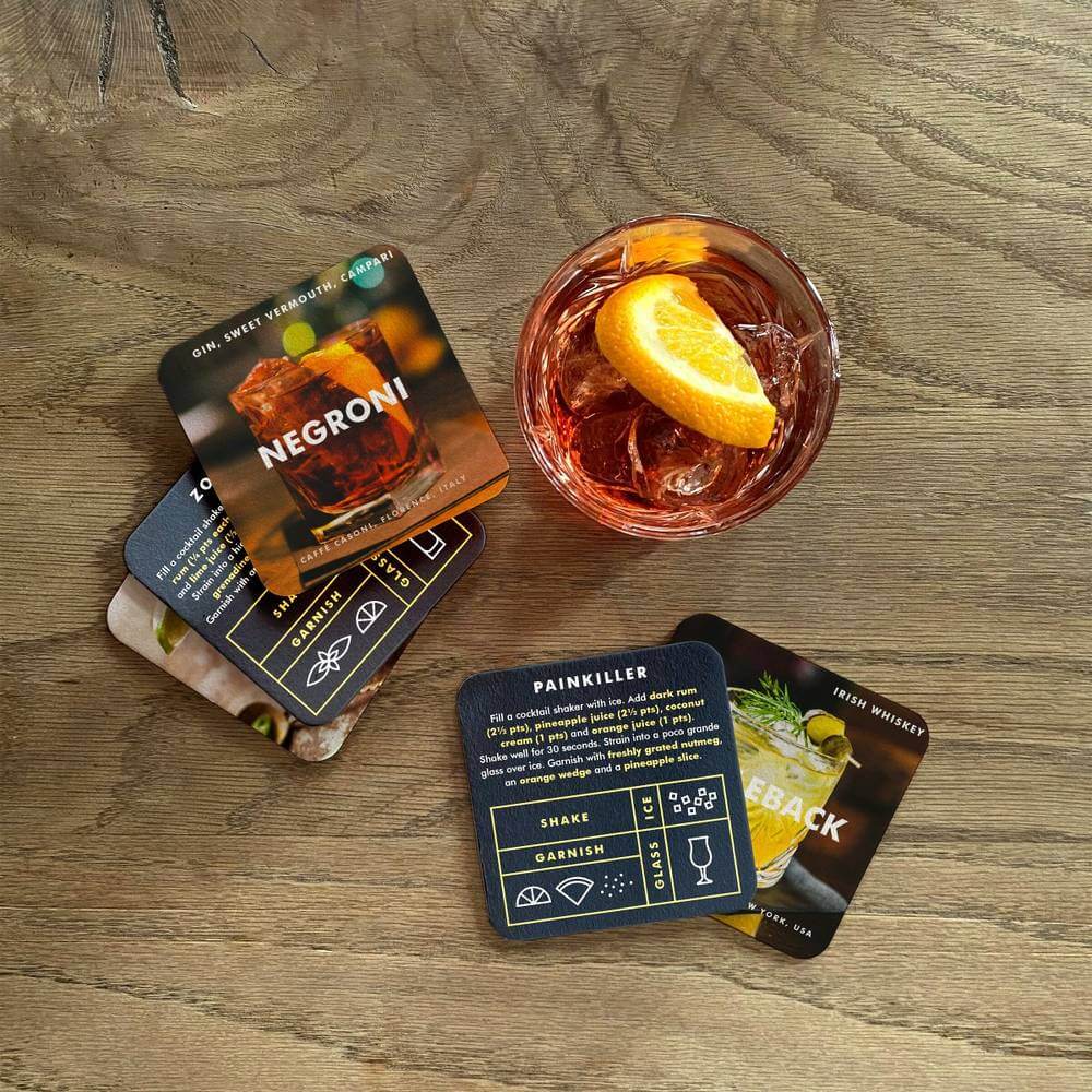 Cocktail recipe coasters on wood with a Negroni card and lemon garnish drink.