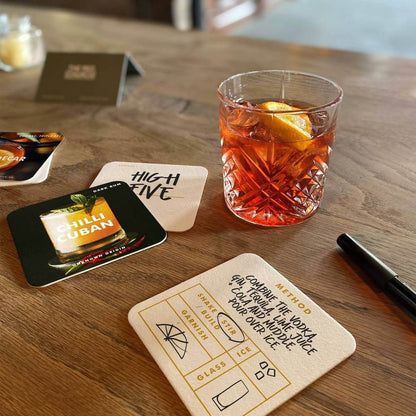 Cocktail recipe coasters on a table with an amber drink in a glass and ice.