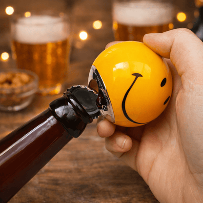 Hand holding yellow smiley pool ball bottle opener beside a brown beer bottle