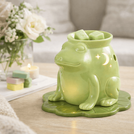 Green frog-shaped wax warmer on a table with candles and flowers in the background