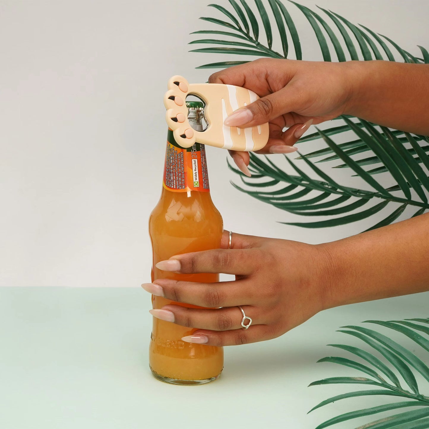 Hands using a beige cat bottle opener magnet to remove a bottle cap from a drink bottle.