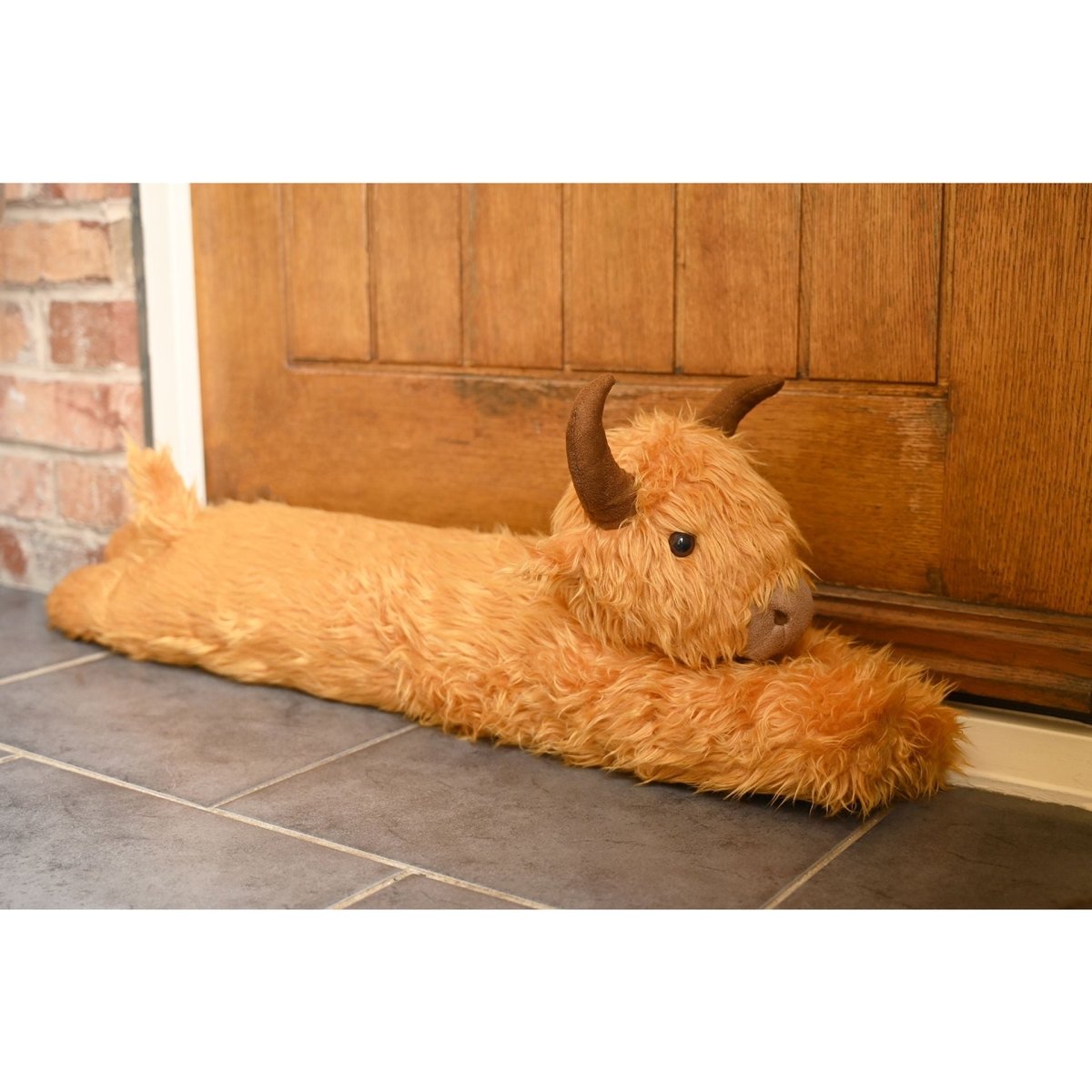 Orange plush Highland cow draught excluder lying on floor tiles, head facing forward.
