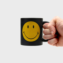 Black ceramic mug with yellow smiley face; hand grips the right-side handle