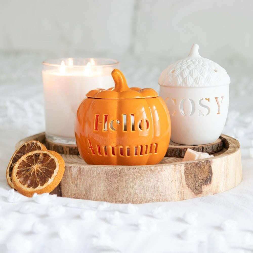 Orange pumpkin wax melt burner on wood base with lit candle and white decorative jar
