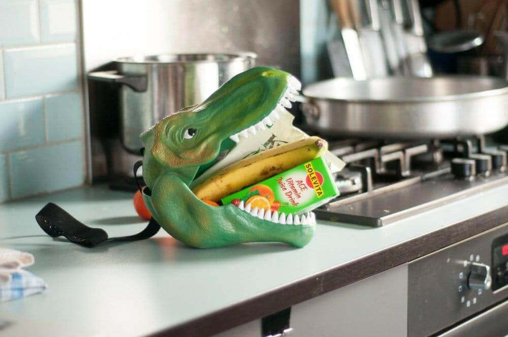 Green T-rex lunch box with black strap on a kitchen counter, snack visible