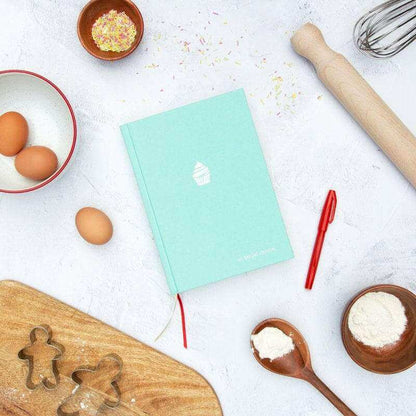Turquoise baking journal book with white cupcake icon, red ribbon, and baking accessories