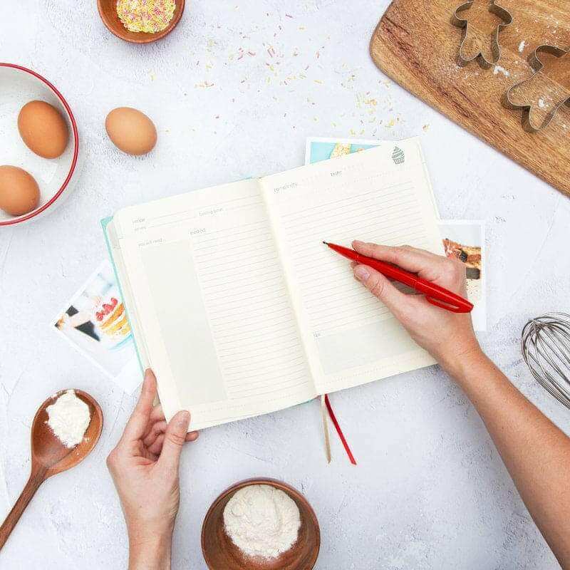 Open baking journal book with lined pages and a hand holding a red pen