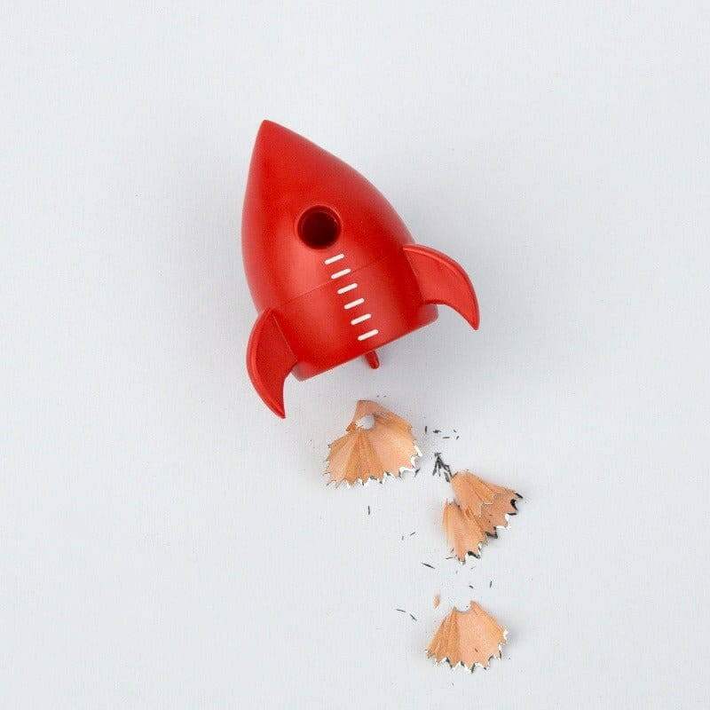 Red rocket pencil sharpener releasing wood shavings; rocket shape with side fins.