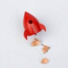 Red rocket pencil sharpener releasing wood shavings; rocket shape with side fins.